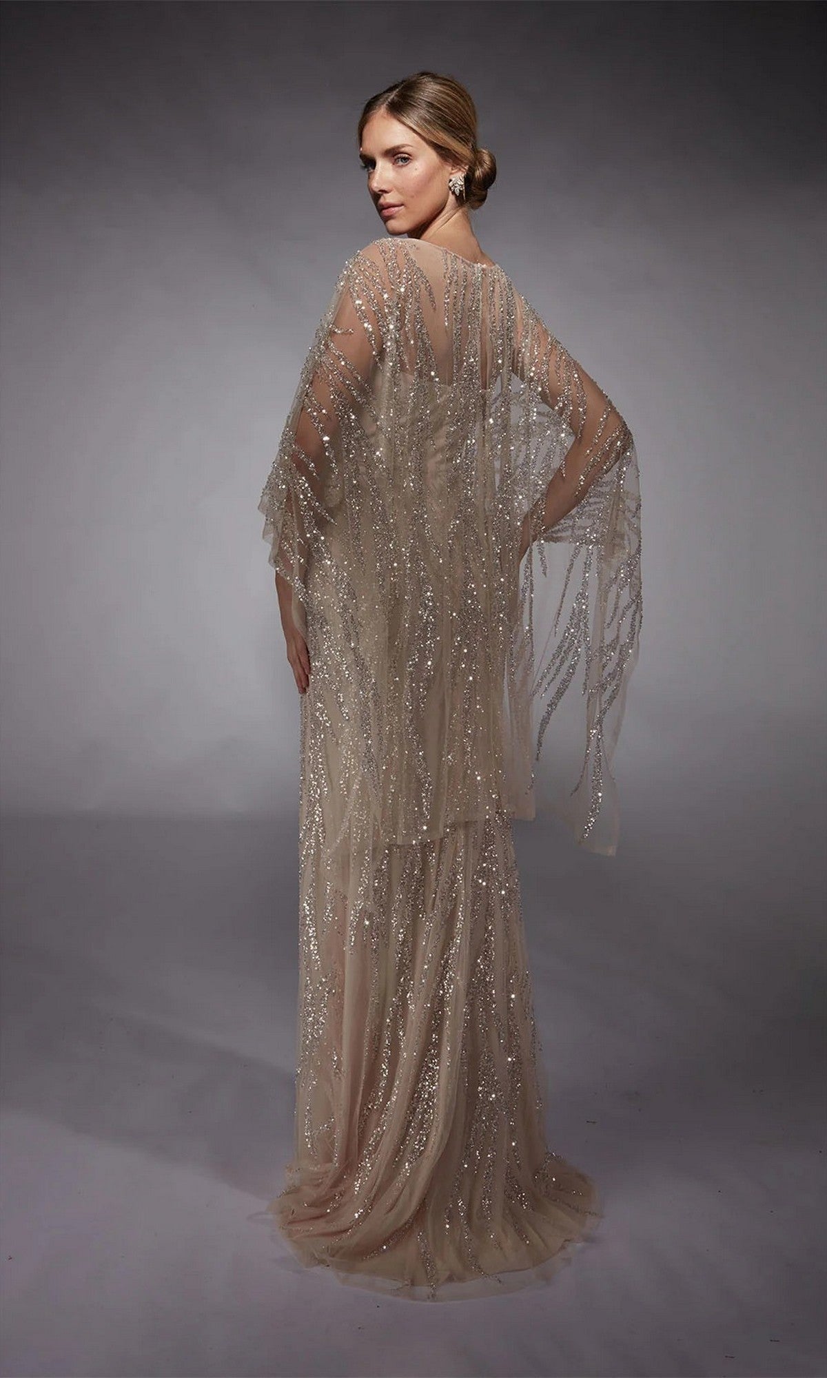 A woman models the Alyce 27754, a floor-length sheer sequin gown with sparkling bead embellishments and a low bun, set against a gray background—an elegant formal or mother-of-the-bride dress.